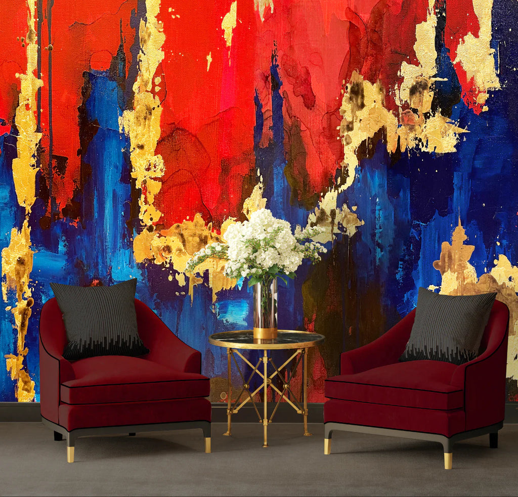 This photo shows a living room full of reds, blues and golds. The foreground features red velvet chairs and black pillows sit beside a glass coffee table with white flower. This accents the large abstract wallpaper design with red and blue tones, gold leaf textures and inky bold shapes.