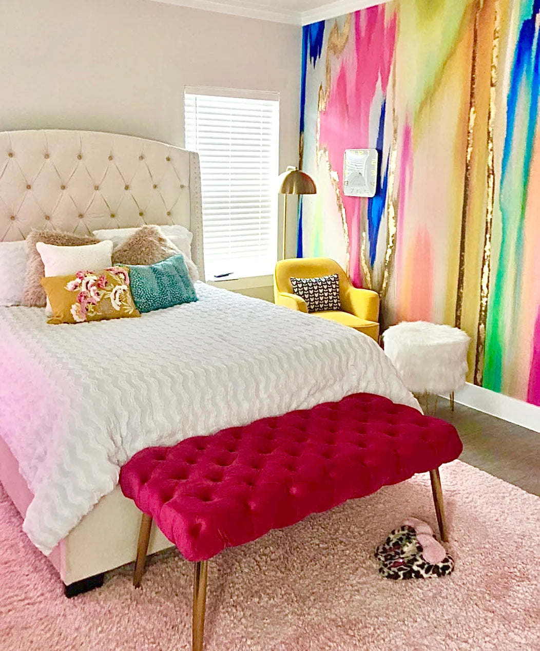 The image displays a kids bedroom with luxury abstract wallpaper. The home decor wallpaper contains fuchsias, blues, mints and yellows along with many other unique colors. This photo also contains and queen bed, tufted chair, yellow love seat, white ottoman and gold lamp.
