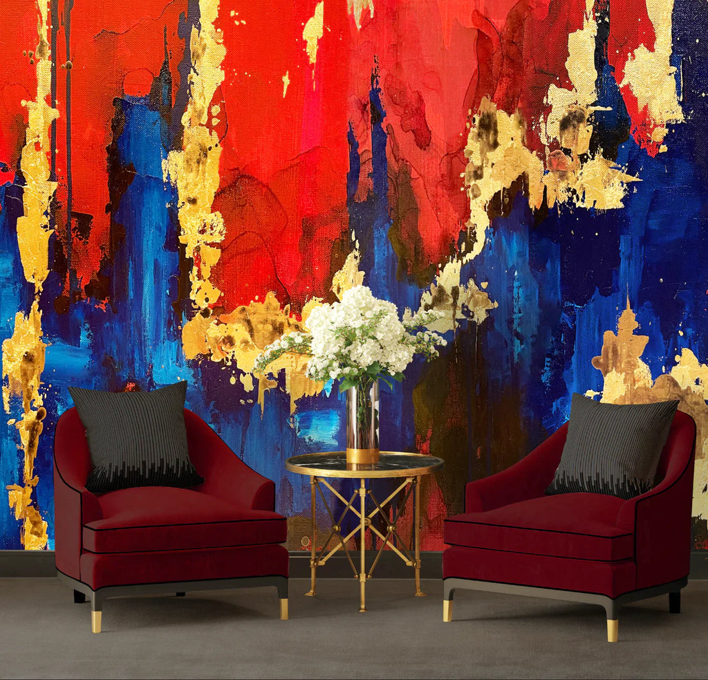 This photo shows a living room full of reds, blues and golds. The foreground features red velvet chairs and black pillows sit beside a glass coffee table with white flower. This accents the large abstract wallpaper design with red and blue tones, gold leaf textures and inky bold shapes.