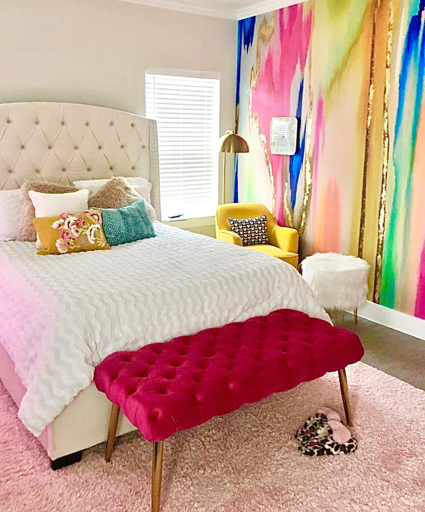The image displays a kids bedroom with luxury abstract wallpaper. The home decor wallpaper contains fuchsias, blues, mints and yellows along with many other unique colors. This photo also contains and queen bed, tufted chair, yellow love seat, white ottoman and gold lamp.  