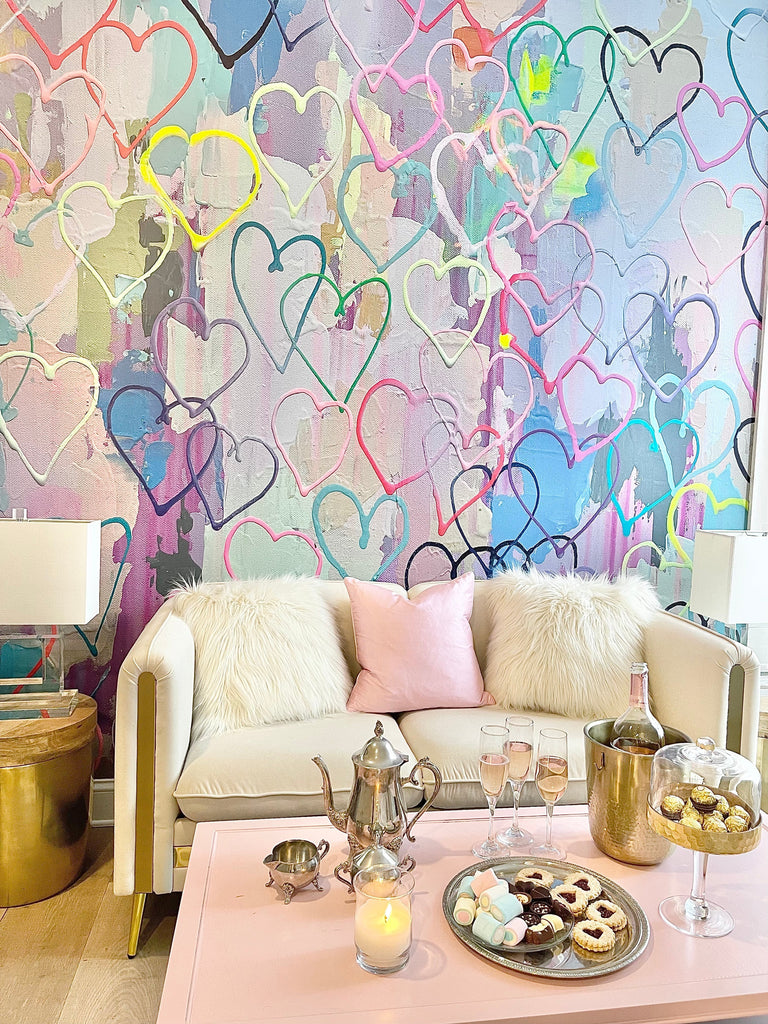 Playful, colorful heart themed wallpaper behind a white sofa, pink coffee table, candles, champagne. This wallpaper is available in prepasted and peel and stick materials. The design is a digitally recreated original painting.