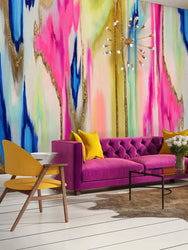This product photo displays a fully staged concept for our best home interior wallpaper design, El Dorado. This design is vibrant with fuchsias, blues, mints, golds, yellows and many more unique colors. This abstract wallpaper design has been featured in homes, hotels, airbnbs, offices, spas and salons. This product photo shows a fuchsia luxury couch, wooden and yellow cloth chairs, glass tables, floral arrangements, and yellow pillows.