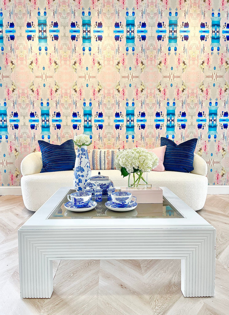 This modern living room showcases a large pattern interior wallpaper design with lush beiges, pinks and blues. The rounded back sofa and blue pillows compliment the wall colors perfectly. The foreground of this interior design staging has a white coffee table and fine China plates and cups.
