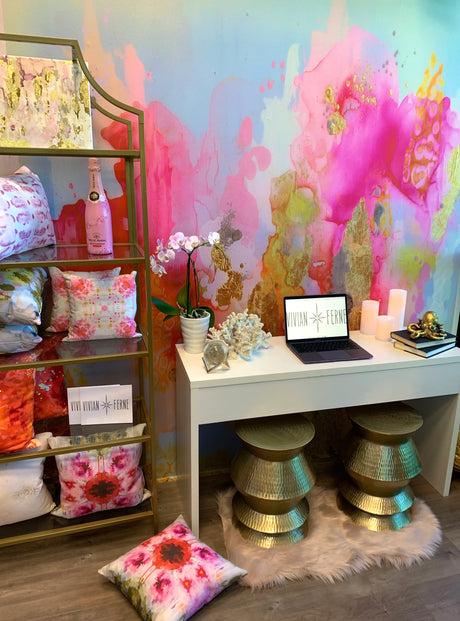 Fully staged interior design space with pink tropical wallpaper. Image also features a white desk, gold stools, gold shelving and multicolor pillows. Other decor elements include books, octopus sculpture, candles and orchids.