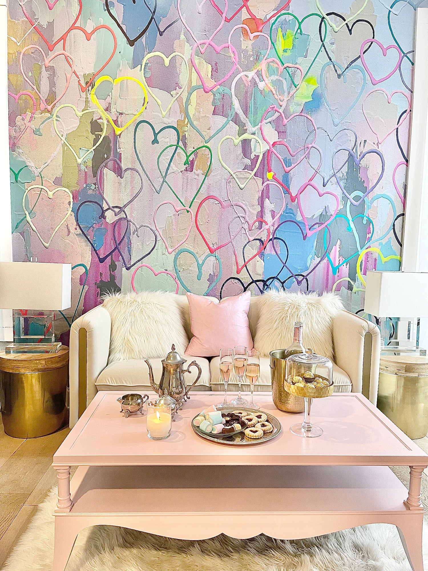 Wallpaper product shoot with multicolored heart themed interior wallpaper decor. This wallpaper design features hand crafted heart shapes, bold acrylic paint textures that make the wallpaper feel like it is jumping off the wall. This wall decor is available in prepasted, peel and stick and french lux materials. The image also features a pink coffee table, champagne glasses, chocolates, tea kettles end tables, and glass lamps.