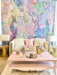 Wallpaper product shoot with multicolored heart themed interior wallpaper decor. This wallpaper design features hand crafted heart shapes, bold acrylic paint textures that make the wallpaper feel like it is jumping off the wall. This wall decor is available in prepasted, peel and stick and french lux materials. The image also features a pink coffee table, champagne glasses, chocolates, tea kettles end tables, and glass lamps.